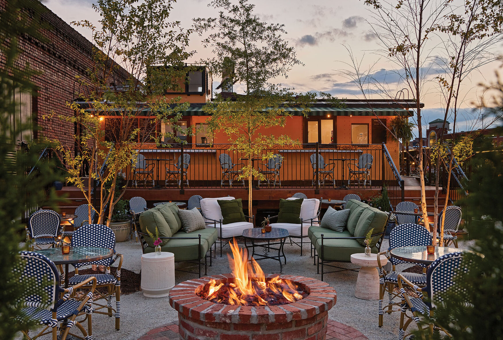 Patio furniture around a circular brick fire pit outside of an orange trailer.